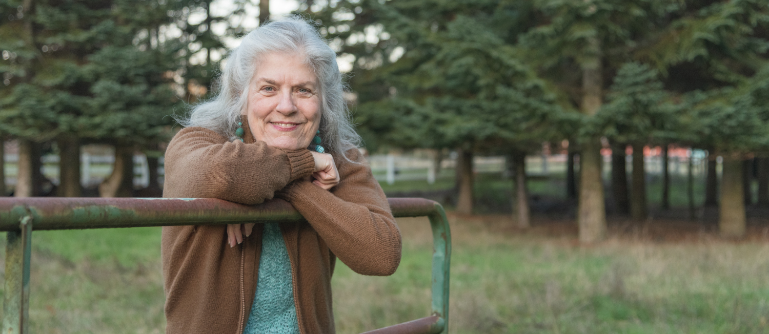 Photo of author's mother, Debra Bond-Yancey: A smiling woman with shoulder-length gray hair leaning on an old green gate in front of green fir trees