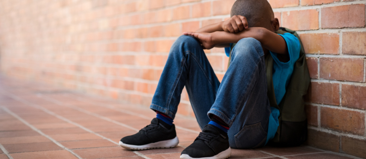 Photo of young Black child with short hair sitting on the ground with their head in their hands.