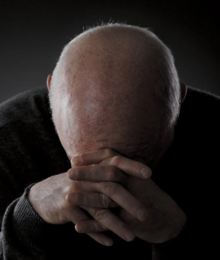 Screen capture of article photo of older human with head down and hands grasping head